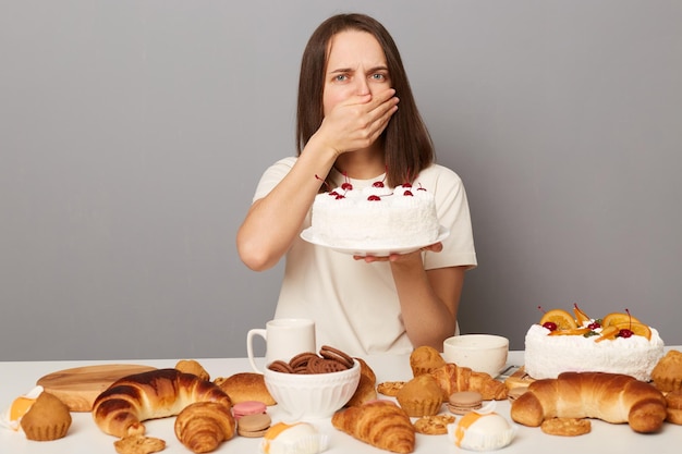 Retrato de una mujer enferma de cabello castaño con camiseta blanca sentada en la mesa y siente náuseas de los dulces cubriendo la boca con la mano sosteniendo un pastel aislado sobre un fondo gris