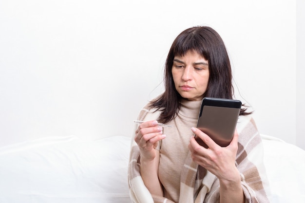 Retrato de una mujer enferma cubierta con una manta sosteniendo un termómetro y comunicándose con el médico en la tableta.