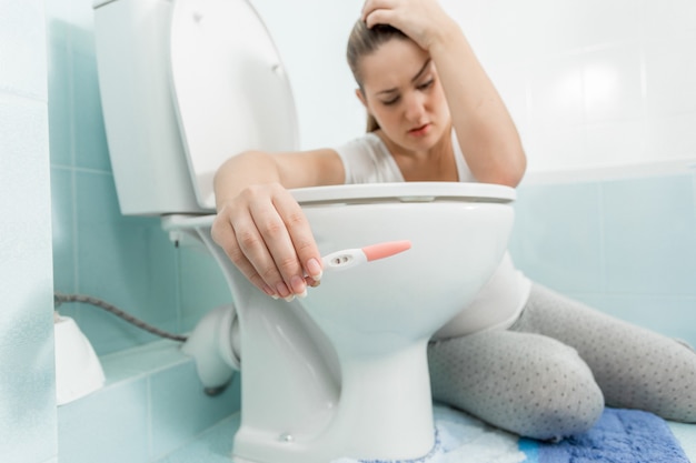 Retrato de mujer enferma triste sentada en el baño y sosteniendo la prueba de embarazo positiva