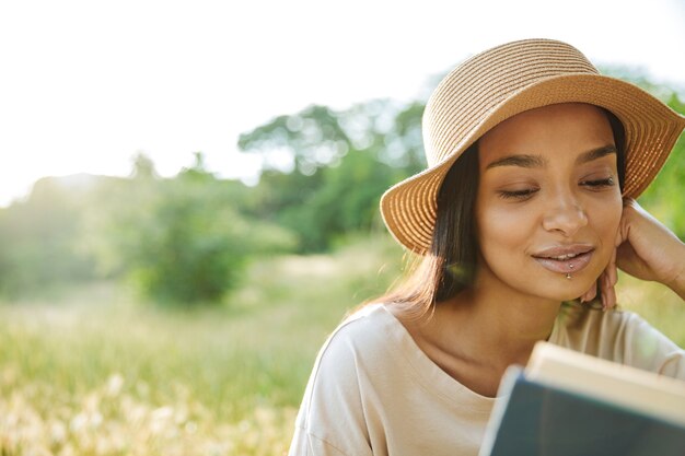 Retrato de mujer enfocada con piercing de labios y libro de lectura de sombrero de paja mientras está sentado sobre el césped en el parque verde