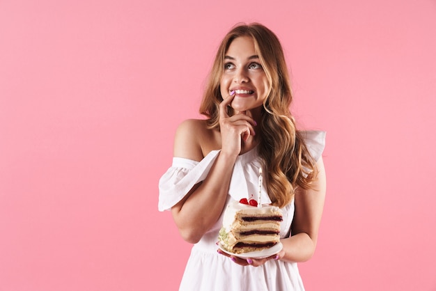 Retrato de mujer de ensueño atractiva con vestido blanco sosteniendo un trozo de pastel con vela aislado sobre pared rosa