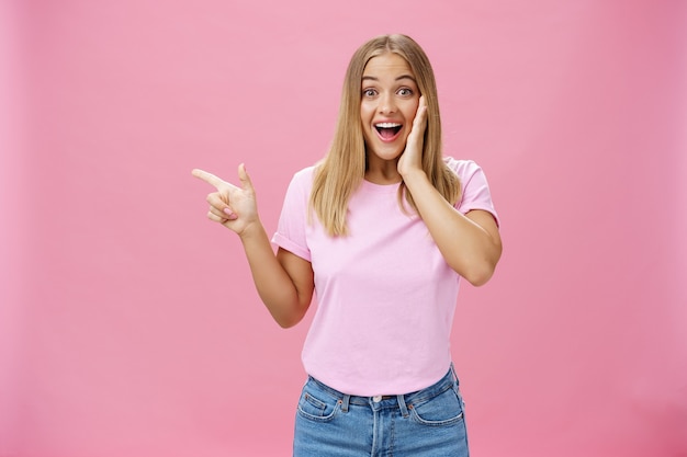 Retrato de mujer entusiasta sorprendida toca su mejilla y señala a la izquierda encantada y divertida sobre fondo rosa.