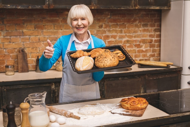 El retrato de la mujer envejecida mayor feliz sonriente atractiva está cocinando en cocina. Abuela haciendo sabrosa hornada. Pulgares hacia arriba.