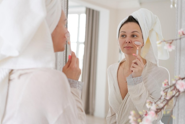 Retrato mujer envuelta en toalla blanca y bata de baño mira el reflejo en el espejo para masajear su cara con rodillo de bola en forma de Y Concepto de cosmética natural alisado de arrugas Enfoque selectivo