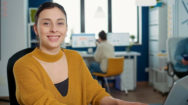 Foto retrato de una mujer escribiendo con el teclado de un portátil y sonriendo a la cámara sentada en el escritorio en una oficina de inicio ocupada. empleado de negocios informal que trabaja relajado con papeles con gráficos y estadísticas.