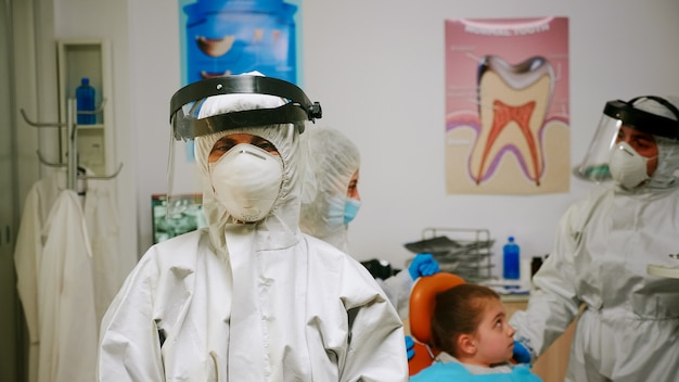 Retrato de mujer estomatóloga cansada con mono y protector facial mirando a la cámara sentado en el nuevo consultorio dental normal. Enfermera pediátrica hablando con el paciente infantil en segundo plano.