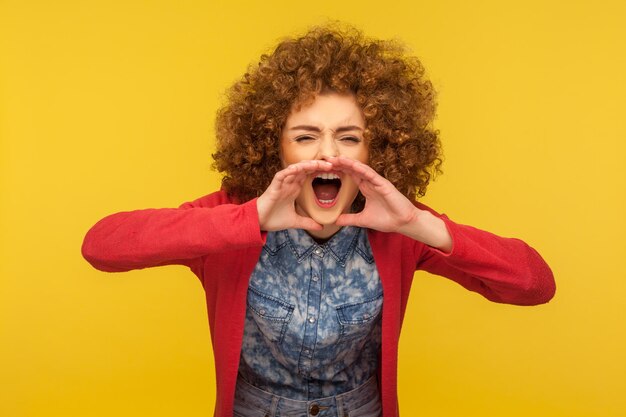 Foto retrato de una mujer estresada con el pelo rizado y ropa informal gritando fuerte en las manos con forma de megáfono, gritando por ayuda, anunciando un mensaje importante. foto de estudio interior, fondo amarillo