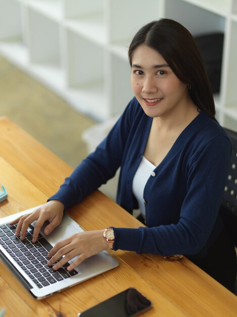 Retrato de mujer estudiante universitaria sonriendo a la cámara mientras realiza una tarea con un portátil en la biblioteca