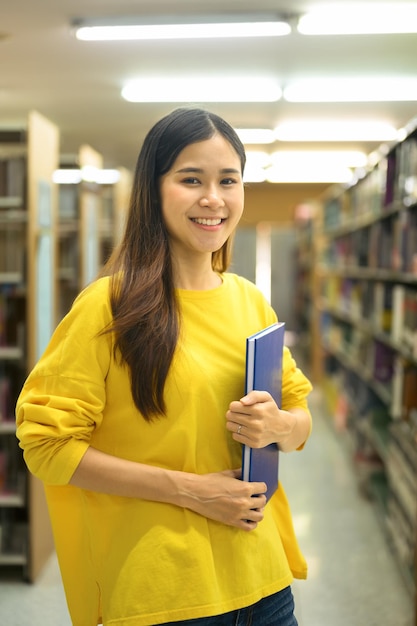 Retrato de mujer estudiante universitaria sosteniendo libros de texto de pie en la biblioteca mirando a la cámara Concepto de conocimiento y educación