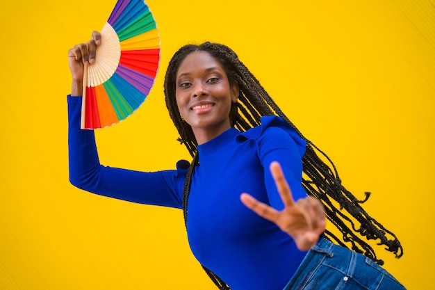 Retrato de una mujer de etnia negra bailando con un abanico lgbt de arco iris sobre un fondo amarillo