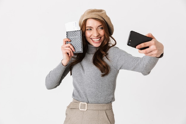 Retrato de mujer europea de 30 años con sombrero tomando selfie en teléfono celular mientras sostiene pasaporte y boletos de viaje aislados en blanco