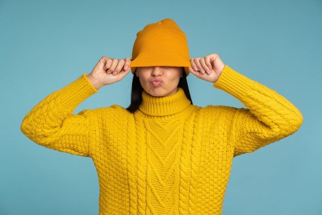 Retrato de mujer europea complacida con suéter de punto tocando su sombrero con las manos y mostrando caras divertidas mientras disfruta del invierno aislado en azul