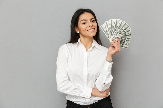 Retrato de mujer exitosa sonriente con cabello largo castaño vistiendo ropa de oficina sonriendo y sosteniendo billetes de un dólar de dinero, aislado sobre fondo gris