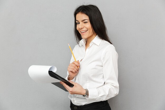 Retrato de mujer exitosa vistiendo traje formal sosteniendo portapapeles con papeles y lápiz para escribir notas en documentos, aislado sobre fondo gris