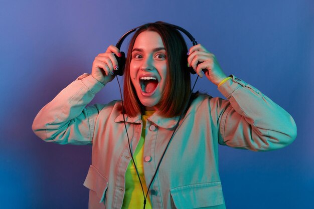 Retrato de una mujer extremadamente feliz y emocionada con una elegante chaqueta posando aislada en un fondo de luz de neón de pie con auriculares y disfrutando del nuevo álbum de su cantante favorito