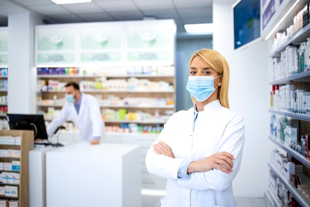 Retrato de mujer farmacéutica con mascarilla y bata blanca de pie en la farmacia durante la pandemia del virus corona.