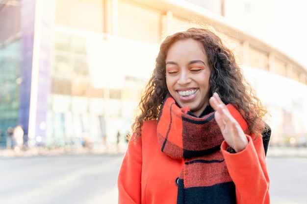 Retrato de una mujer feliz con un abrigo naranja y una bufanda en una ciudad urbana reuniéndose con amigos