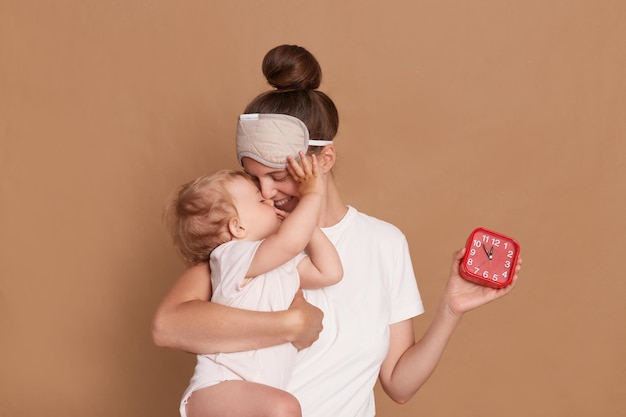 Retrato de una mujer feliz y alegre con el pelo oscuro y los ojos vendados de pie con su hija pequeña y un reloj despertador besando y abrazando a su hijo posando aislado sobre un fondo marrón