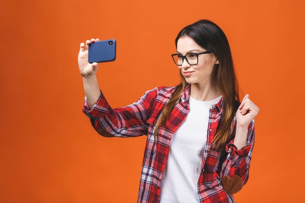 Retrato de la mujer feliz bastante joven que hace el selfie en smartphone, aislado contra fondo anaranjado.