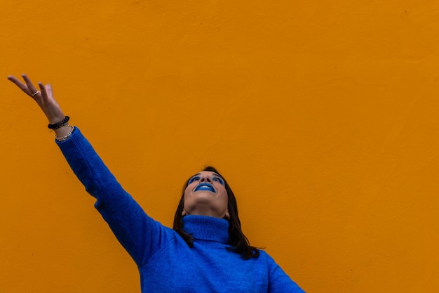 Retrato de una mujer feliz con el brazo levantado y mirando hacia arriba vestida de azul sobre un fondo amarillo