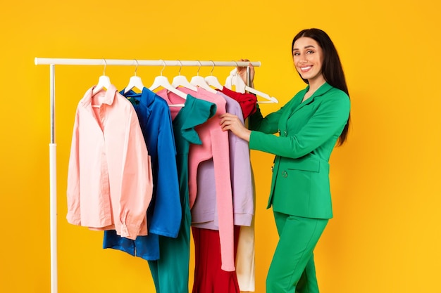 Retrato de una mujer feliz diseñadora de moda de pie cerca de un estante de prendas de vestir, una mujer estilista presentando nueva ropa de moda