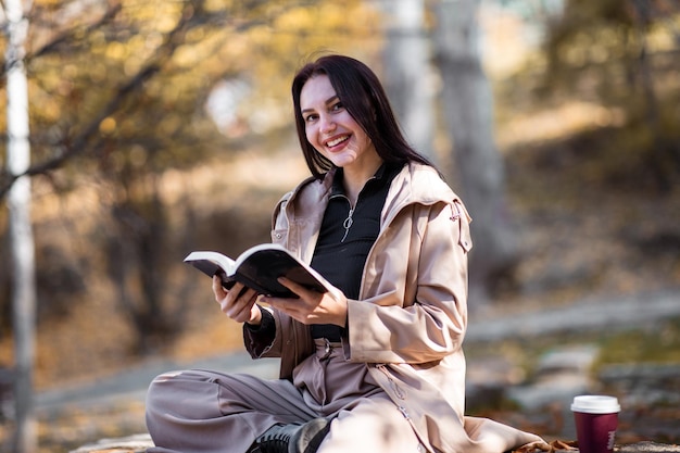 Retrato de mujer feliz y elegante en la temporada de otoño con gabardina, está tomando café y leyendo un libro