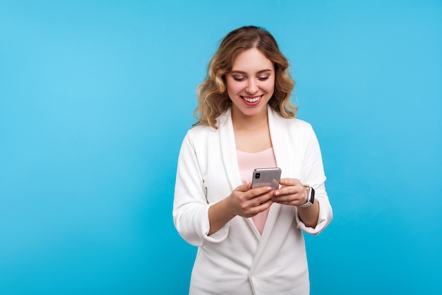 Retrato de una mujer feliz y encantadora con el pelo ondulado en una chaqueta blanca escribiendo un mensaje en el celular y sonriendo, usando aplicaciones móviles, comunicación en línea en las redes sociales. foto de estudio, fondo azul