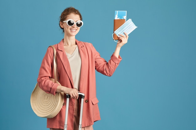 Retrato de mujer feliz en gafas de sol con billetes y pasaporte sonriendo a la cámara de pie contra el fondo azul.