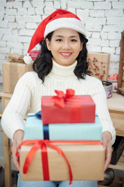 Retrato de mujer feliz con gorro de Papá Noel dando pila de regalos de Navidad y sonriendo a la cámara