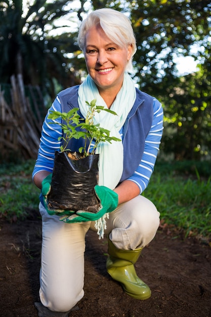 Retrato de mujer feliz jardinero arrodillado con planta en maceta en el jardín