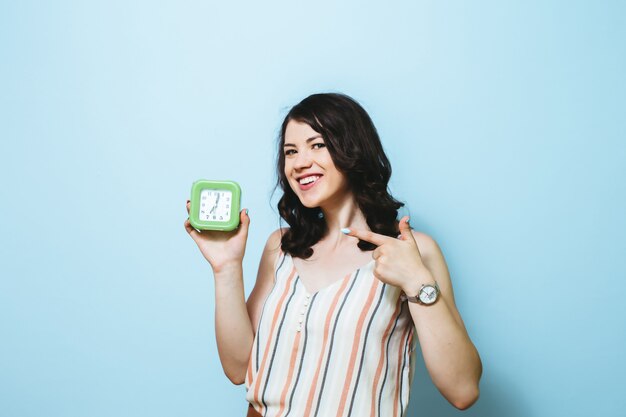 Retrato de la mujer feliz joven aislada sobre la pared azul que sostiene el despertador.