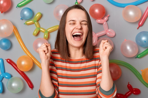 Retrato de una mujer feliz y llena de alegría con el pelo castaño y un vestido a rayas de pie con los puños apretados alrededor de globos inflados multicolores regocijándose gritando