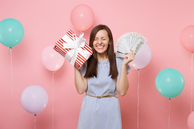 Retrato de mujer feliz llena de alegría en vestido azul sosteniendo un montón de dólares en efectivo y caja roja con regalo presente sobre fondo rosa con coloridos globos de aire. Concepto de fiesta de cumpleaños.