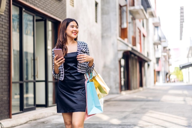 Retrato de mujer feliz de moda asiática de belleza sonriente relajarse y disfrutar del tiempo de compras venta de verano caminar y sostener una bolsa de compras colorida para comprar algo compras usar teléfono inteligente en la tienda