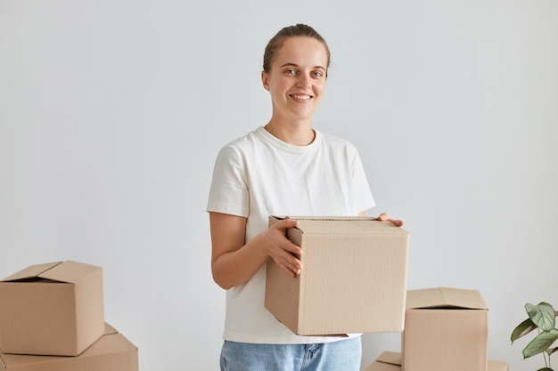 Retrato de una mujer feliz con peinado de moño usando una camiseta blanca de estilo casual, mirando la cámara, de pie con un paquete de cartón con pertenencias, bienes raíces, comprando o alquilando una casa nueva.