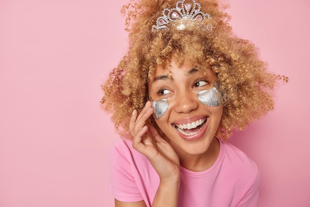 Foto el retrato de una mujer feliz con el pelo rizado y tupido lleva una corona que aplica parches de belleza plateados debajo de los ojos para eliminar las arrugas vestida con una camiseta aislada sobre un espacio de copia de fondo rosa para tu texto