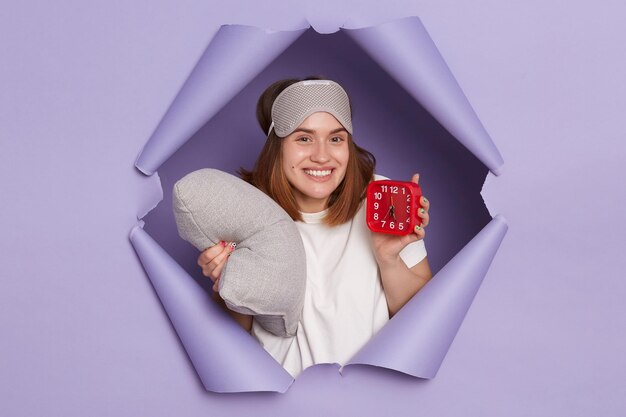 Retrato de una mujer feliz y positiva con camiseta blanca y una venda en los ojos posando en una pared rota de papel morado despertándose de buen humor sosteniendo un despertador y una almohada en las manos