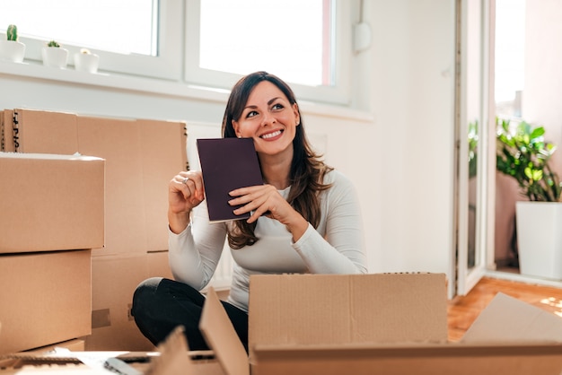 Retrato de una mujer feliz que se sienta en el piso entre las cajas con un libro. Mudarse a un nuevo apartamento.