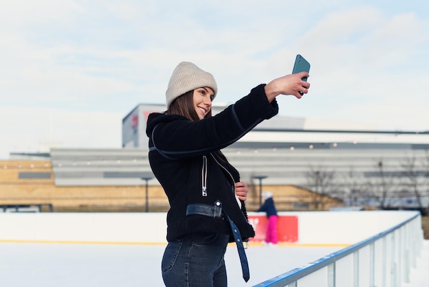 Retrato de una mujer feliz con ropa de invierno haciendo una foto selfie en un smartphone mientras patina en el hielo