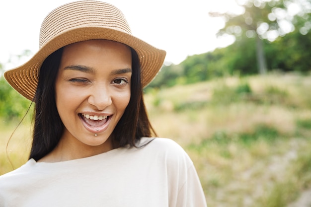 Retrato de mujer feliz con sombrero de paja y piercing de labios guiñando un ojo a la cámara mientras camina en el parque verde