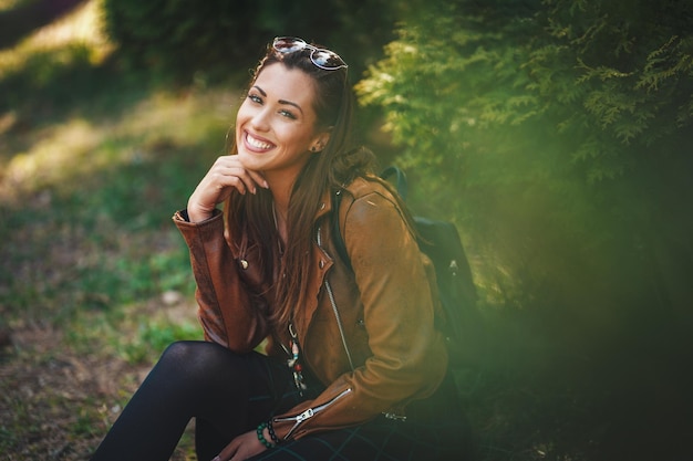 Un retrato de una mujer feliz y sonriente, con una mochila en la espalda, que está sentada en la hierba verde y disfrutando de un día soleado al aire libre.