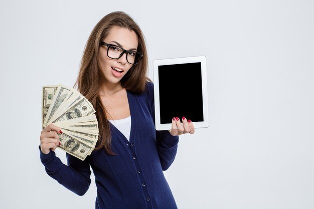 Retrato de una mujer feliz sosteniendo dinero y mostrando la pantalla de la tableta en blanco aislado en un fondo blanco.