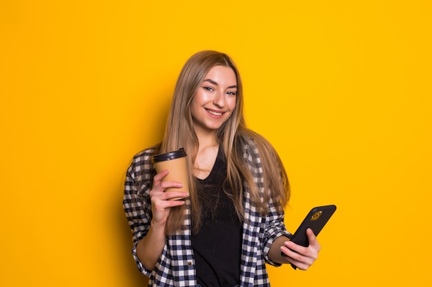 Retrato de una mujer feliz sosteniendo la taza de café mientras está de pie y usando el teléfono móvil en una pared amarilla