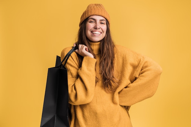 Retrato de mujer feliz con suéter de estilo casual, de pie y sosteniendo bolsas de la compra, mirando a la cámara con una sonrisa, satisfecho con la compra. Tiro de estudio interior aislado sobre fondo amarillo