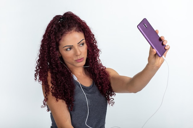 Retrato de una mujer feliz con teléfono móvil y auriculares, sonriendo y haciendo un selfie. Aislado en un fondo blanco.