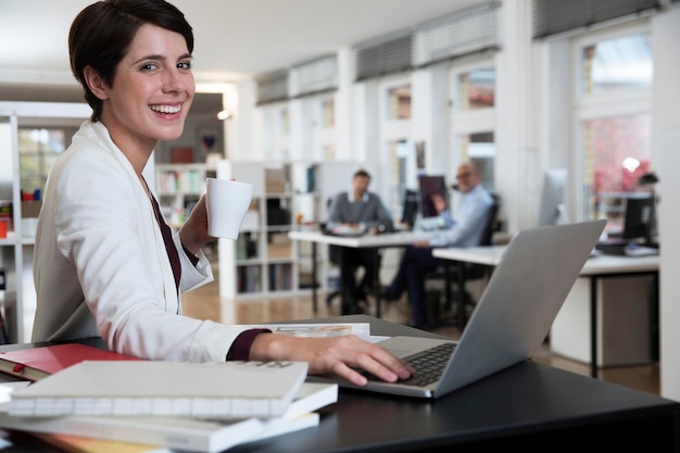 Retrato de mujer feliz usando un portátil en la oficina con colegas en segundo plano.