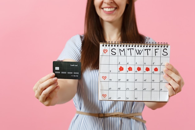 Retrato de mujer feliz en vestido azul, sombrero con tarjeta de crédito, calendario de períodos, comprobación de días de menstruación aislados sobre fondo rosa de tendencia. Concepto ginecológico de atención médica. Copie el espacio.