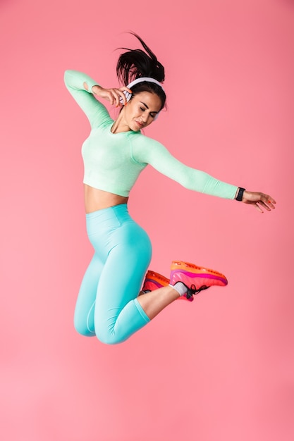 Retrato de mujer de fitness deportivo joven hermosa emocional aislada sobre pared rosa pared saltando escuchando música con auriculares.