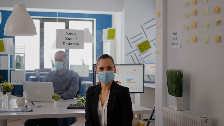 Foto retrato de mujer gerente con mascarilla para prevenir la infección por coronavirus sentada en una silla en la mesa de escritorio en la oficina de negocios. los compañeros mantienen el distanciamiento social utilizando una placa de plástico separada