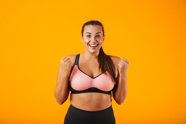 Retrato de mujer gordita feliz en sujetador deportivo sonriendo y de pie, aislado sobre fondo amarillo